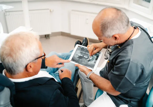 Zahnarzt erklärt einem Patienten mithilfe eines digitalen Röntgenbilds die Notwendigkeit eines Knochenaufbaus vor dem Einsetzen eines Zahnimplantats.