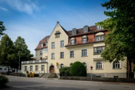 Außenansicht der Zahnarztpraxis Dr. Bayer & Kollegen in der Von-Kühlmann-Straße 1 in Landsberg. Das Gebäude hat eine gelbe Fassade und ein rotes Satteldach. Eine Rampe führt zum Eingang, und es sind zwei Büsche vor dem Gebäude zu sehen.