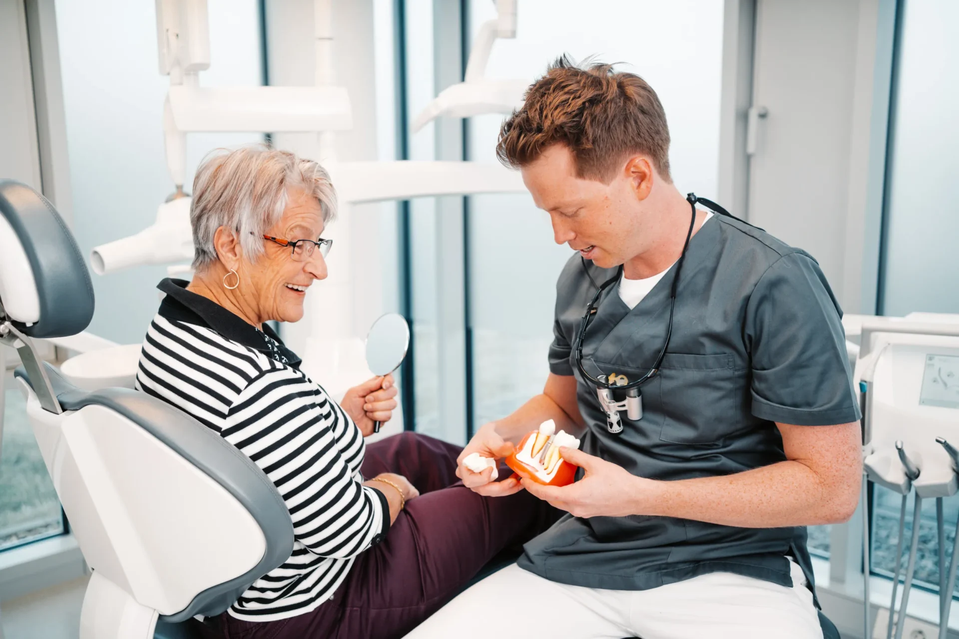 Zahnarzt erklärt einer älteren Patientin mit Zahnmodell und Spiegel das Konzept fester Zähne an einem Tag.