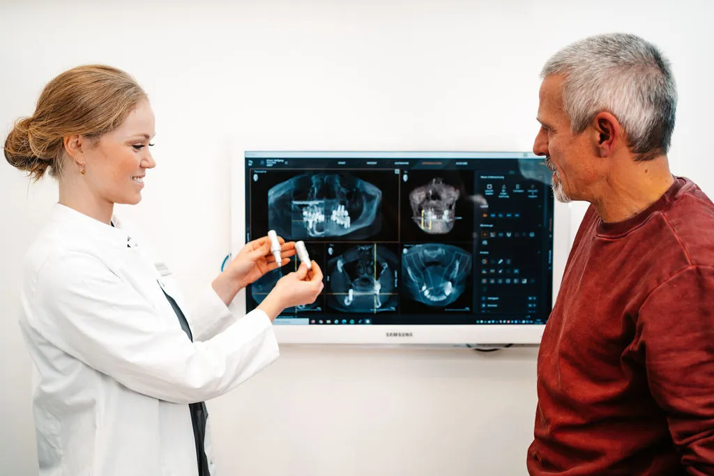 Zahnarzt zeigt einem Patienten digitale Röntgenbilder auf einem Bildschirm und erklärt die Behandlung mit einem Modell.