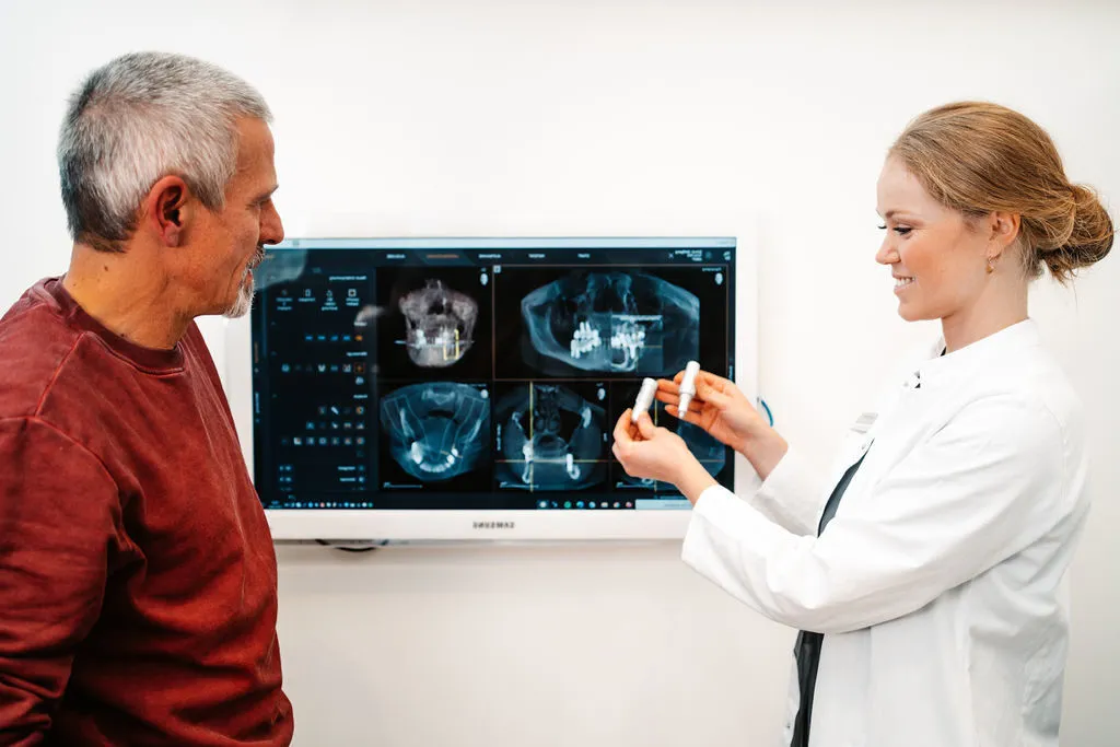 Zahnarzt zeigt einem Patienten digitale Röntgenbilder auf einem Bildschirm und erklärt die Behandlung mit einem Modell.