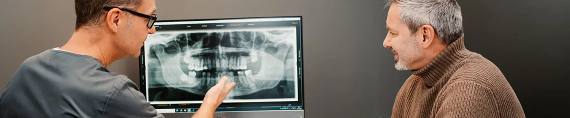 Zahnarzt bespricht mit einem Patienten die Befunde eines digitalen Röntgenbildes auf einem Monitor im Rahmen der Implantatberatung