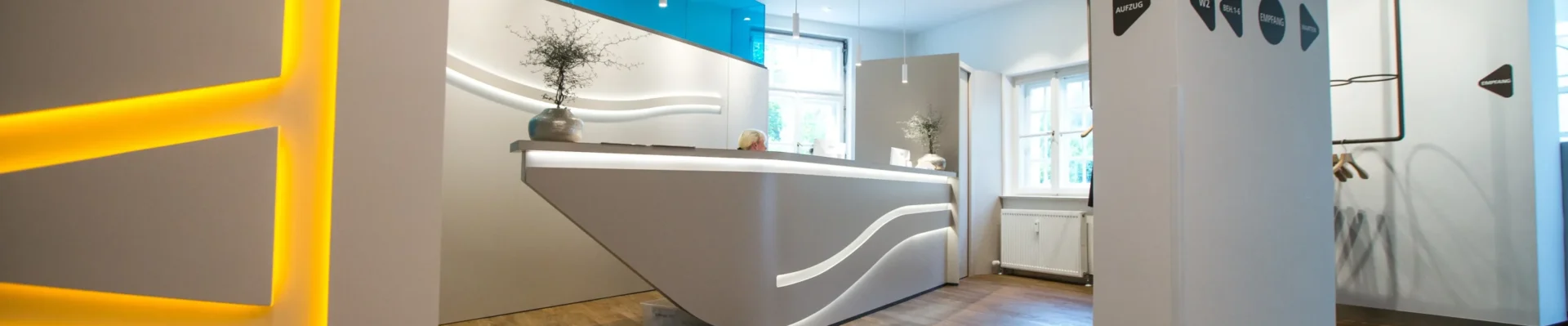 Moderner Empfangsbereich der Zahnarztpraxis in Landsberg mit stilvoller Beleuchtung, Holzfußboden und elegantem Tresen.