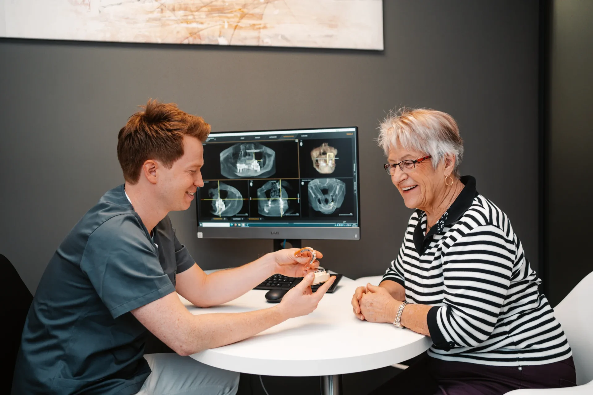 Zahnarzt zeigt einer älteren Patientin eine implantatgestütze Prothese und erklärt die Behandlung.
