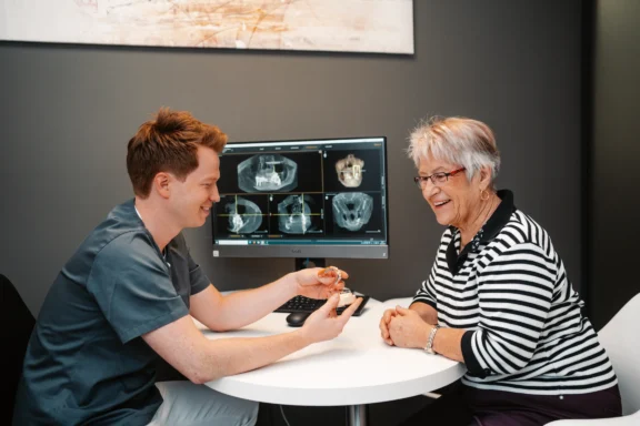 Zahnarzt zeigt einer älteren Patientin eine implantatgestütze Prothese und erklärt die Behandlung.