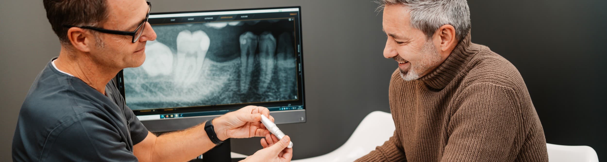 Zahnarzt erklärt einem Patienten den Ablauf einer Implantatbehandlung anhand eines Implantatmodells, im Hintergrund ein digitales Röntgenbild.